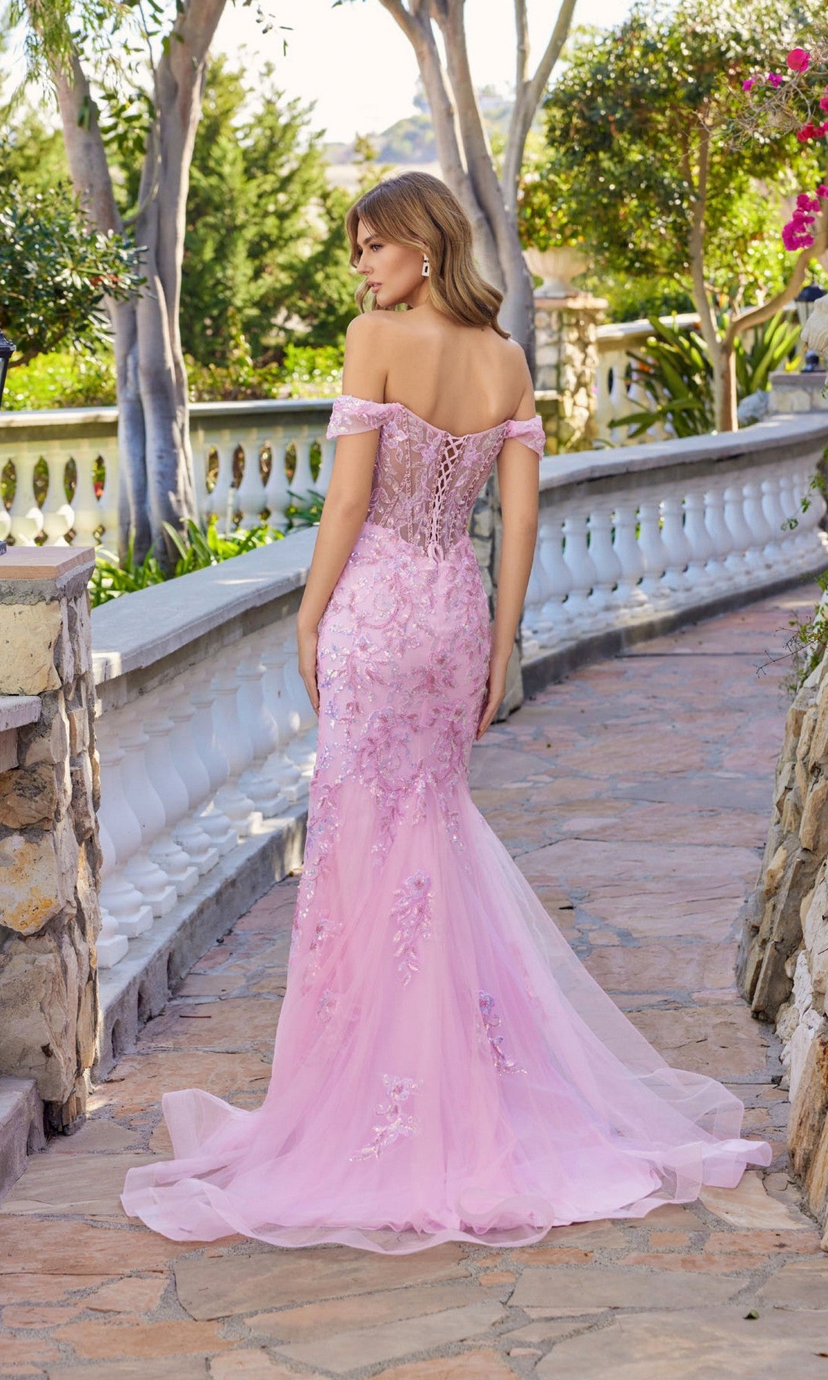 A woman in the Juliet JT2612N long prom dress, featuring a light pink off-the-shoulder mermaid silhouette with floral applique, stands on a stone path bordered by greenery and white railings, facing away from the camera.